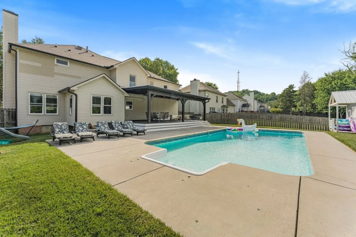 The image highlights a serene backyard setting with a swimming pool, patio seating, a lush green lawn, and a covered porch. The space is designed for both relaxation and lively outdoor dining, offering ample room for family and friends to gather and enjoy meals in the fresh air.
