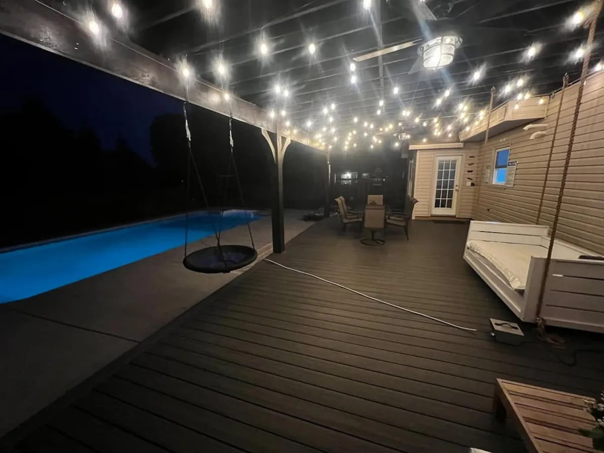 A tranquil outdoor setting illuminated by hanging string lights, featuring a swimming pool, a swing seat, and comfortable deck furniture. The scene evokes evening relaxation and social gatherings by the pool, with ample space for lounging and enjoying the outdoors.