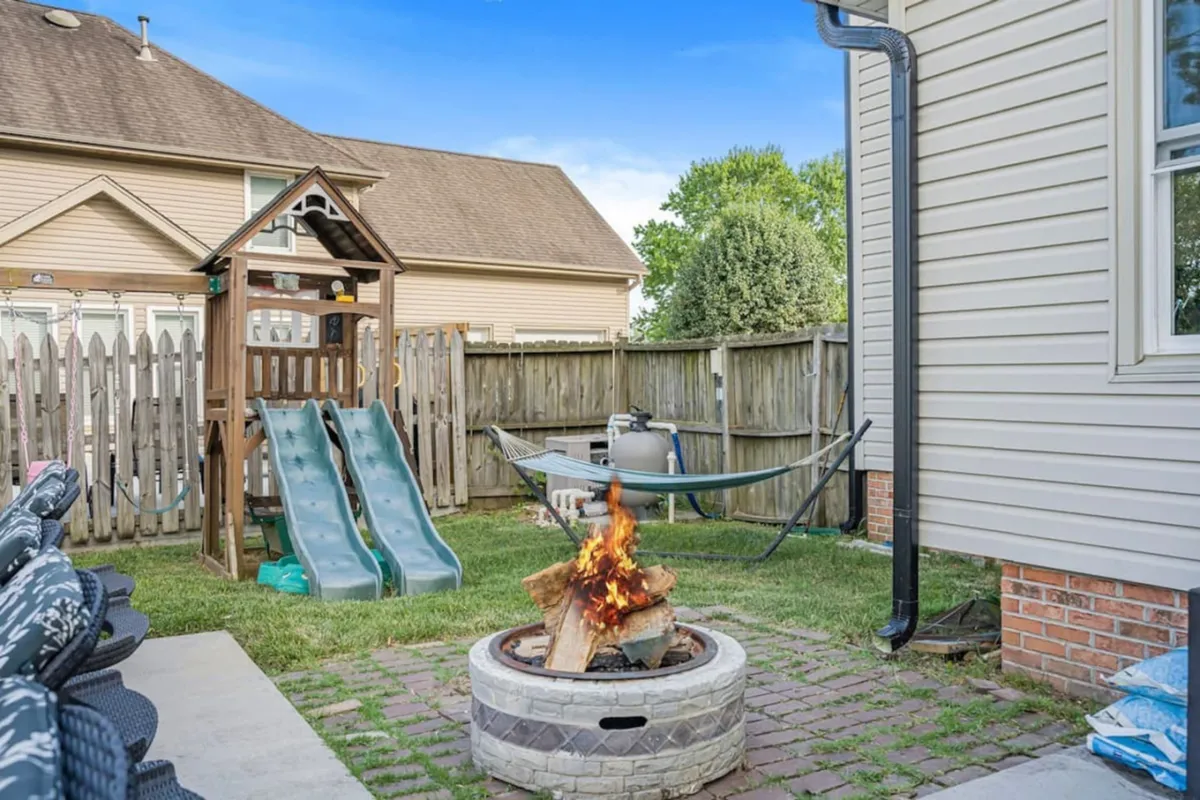 A vibrant, playful backyard featuring a fire pit, hammock, and a children’s playset with slides. The space is bordered by a wooden fence and set up for outdoor family gatherings, playtime, and relaxation.