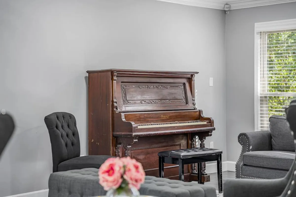 A warmly lit living room centers around a classic piano with a black stool and a vase of fresh flowers atop. The piano is positioned invitingly, suggesting moments of music, relaxation, or family singalongs. The space feels personal and nostalgic, evoking the joy of making music at home.