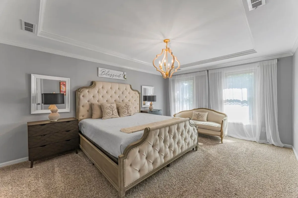 This image presents an elegantly upholstered bed with a chic chandelier and a stylish side table. The scene is tranquil and luxurious, with sunlight streaming in, creating an inviting, restful atmosphere.