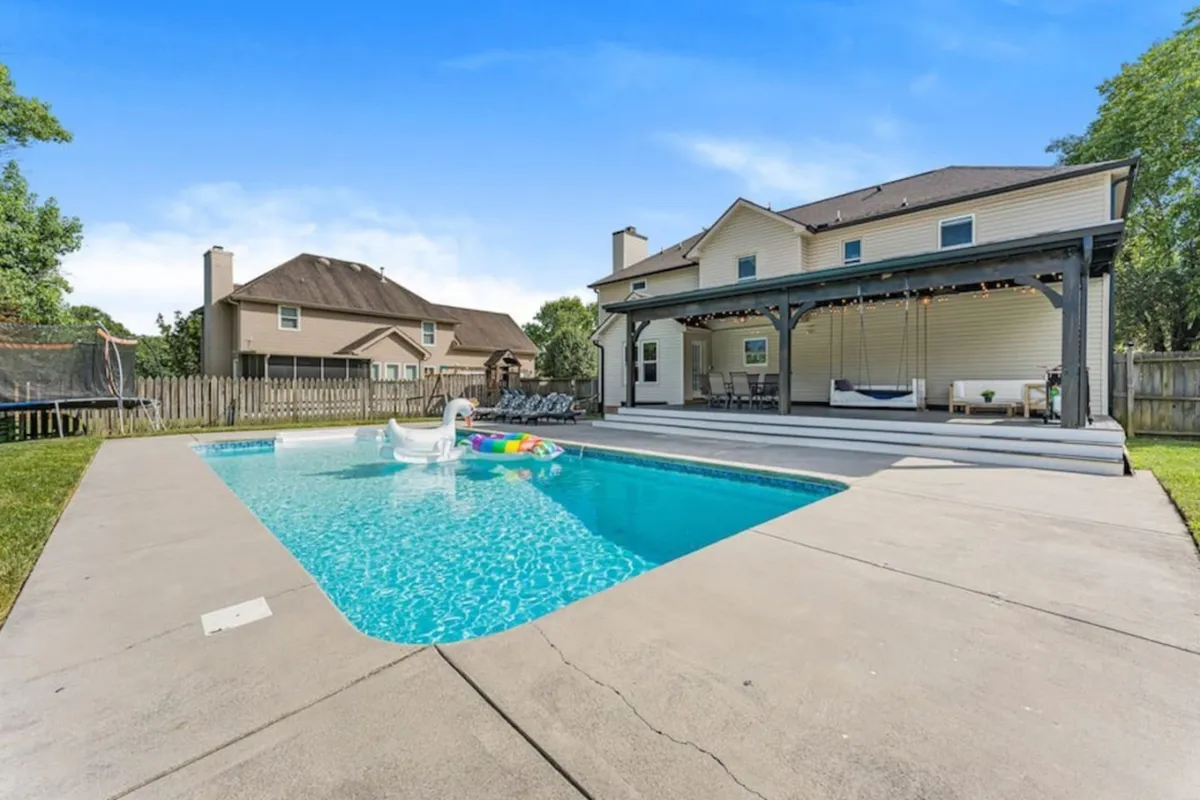 A broad view of the backyard showcasing the swimming pool, ample outdoor seating, the house's facade, and a trampoline nestled nearby. The setup emphasizes a space for both fun and relaxation, with playful and lounging possibilities side by side.