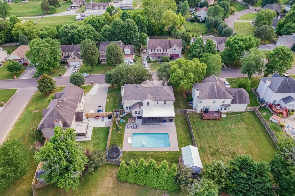 The image showcases a sunlit backyard with a sparkling swimming pool as the focal point, surrounded by lush trees and well-maintained lawns. The inviting outdoor space hints at areas for barbecuing, family games, and relaxed gatherings. The house's exterior is visible, setting the suburban, family-friendly tone.