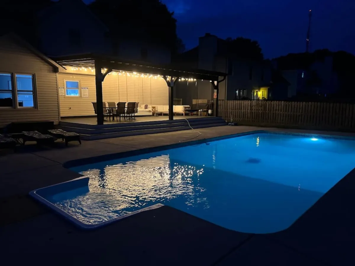 A serene backyard scene featuring a sparkling pool, a covered patio area illuminated by string lights, and ample space for lounging or dining. The atmosphere is tranquil and inviting, ideal for evening relaxation and family fun.