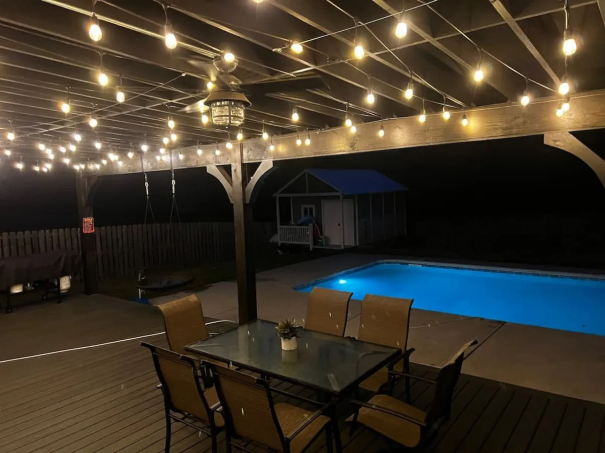 This image features a welcoming outdoor dining area beside a sparkling swimming pool, illuminated by string lights overhead. A dining table with chairs sits on the poolside deck, complemented by a lantern light fixture, evoking a warm and inviting ambiance for evening gatherings or casual meals under the stars.