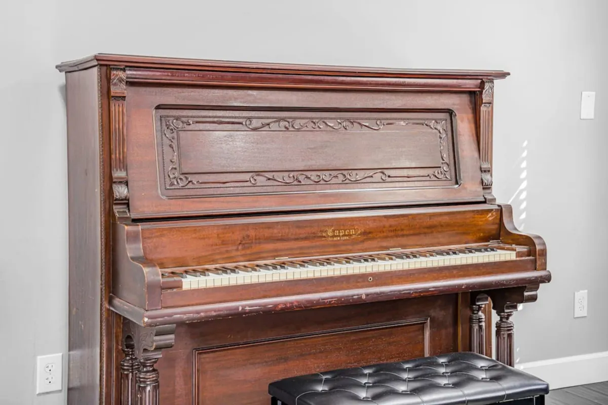 An antique piano with a black bench sits in a quiet, inviting corner. The space exudes nostalgia, perfect for both practice sessions and hosting intimate music gatherings or lessons. The piano's well-loved appearance underscores a tradition of music at home.
