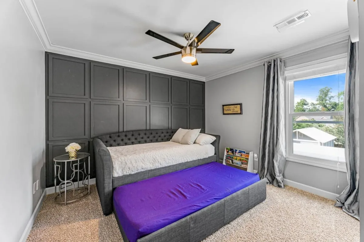 This bedroom image features a bed with a patterned headboard, a daybed with a pull-out mattress, decorative wall paneling, and abundant natural light from a window. The space feels comfortable and casual, suitable for both relaxing and family sleepovers.