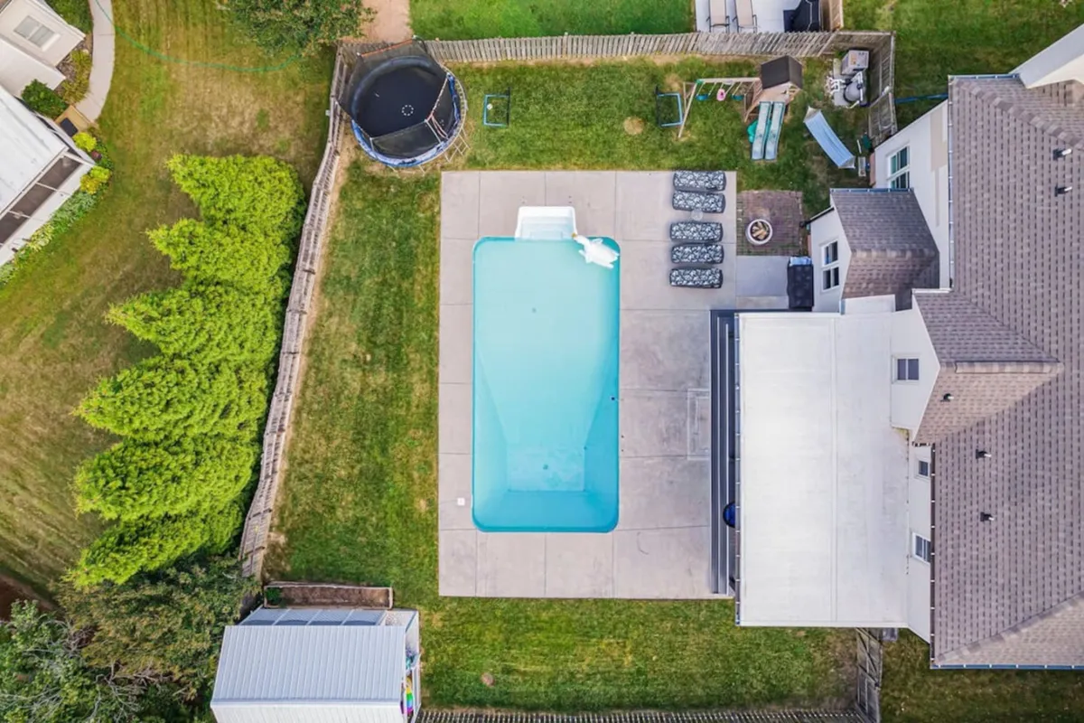A vibrant scene of the backyard featuring a sparkling pool, a trampoline, a play structure, and an outdoor seating area, all set within a lush, inviting environment. The play structure and seating suggest family-friendly fun and gatherings.